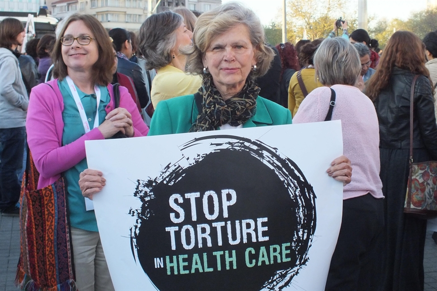 [ai] A woman holding a protest sign that reads "STOP TORTURE IN HEALTH CARE" surrounded by a crowd of people at an event.